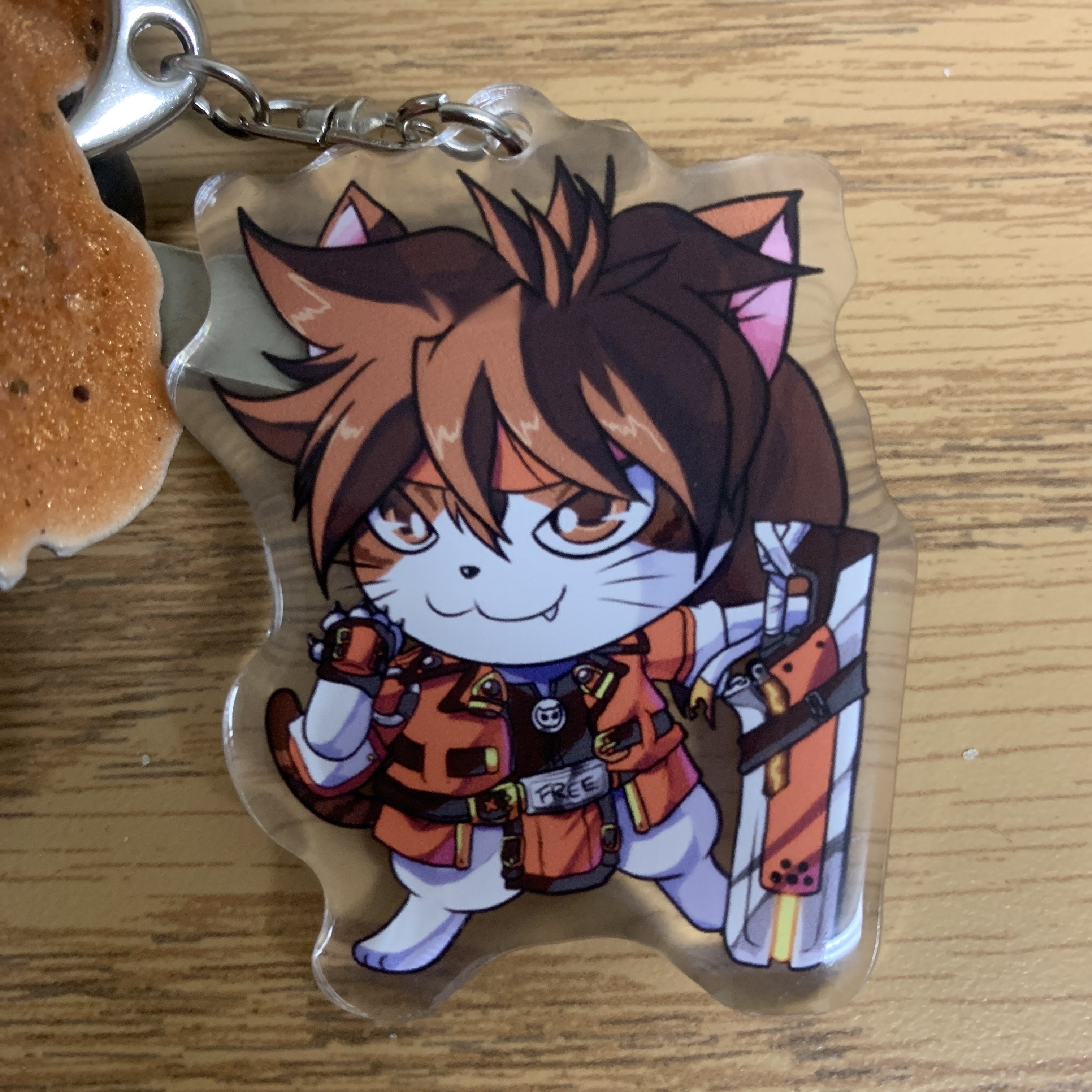 a picture of an acrylic keychain on a desk. it holds art of sol badguy from guilty gear as a chibi anthropomorphic cat. the cat is mostly white with brown on the upper half of its head and its tail and brown eyes, and it is wearing sol's strive outfit and hair